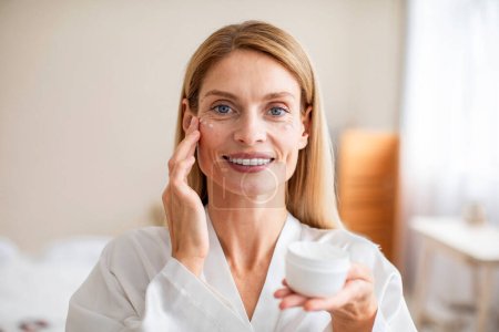 Photo for Beautiful middle aged woman in silky bathrobe using nourishing eye cream, looking at camera and smiling, enjoying face care routine. Dark circles, eye wrinkles treatment - Royalty Free Image