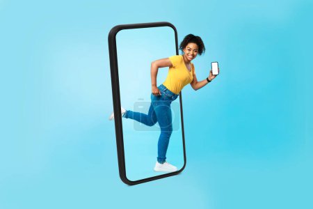 Photo for Smiling funny millennial african american female with smartphone with blank screen runs out of big phone, isolated on blue background, studio. Modern blog, app and gadget for huge sale, ad and offer - Royalty Free Image