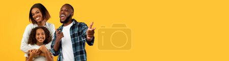 Téléchargez les photos : Heureux père afro-américain, mère et fille posant ensemble sur fond de studio jaune, jolie dame noire étreignant son adolescente, homme joyeux pointant vers la caméra, panorama avec espace de copie - en image libre de droit