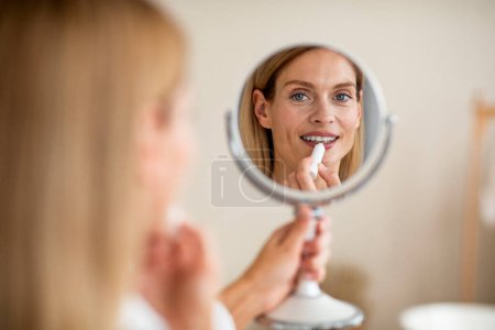 Photo for Nude daily makeup. Attractive middle aged woman applying lipstick on lips, looking at reflection of round mirror in bedroom interior in daylight, copy space - Royalty Free Image