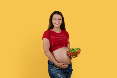 Foto de Concepto de la dieta del embarazo. Mujer embarazada sonriente sosteniendo el tazón con ensalada de verduras y abrazando el vientre, feliz embarazada disfrutando de una nutrición saludable, posando aislada sobre fondo amarillo - Imagen libre de derechos
