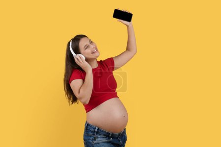 Téléchargez les photos : Joyeuse dame enceinte écoutant de la musique avec smartphone et écouteurs sans fil, Jeune femme enceinte heureuse tenant un téléphone mobile avec écran vide et bénéficiant d'une playlist préférée, Copier l'espace - en image libre de droit