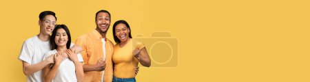 Foto de Romántico joven pareja asiática abrazando y sonriendo, posando junto a afro-americano emocional millennial hombre y mujer apretando puños, celebrando el éxito, amarillo fondo del estudio, espacio de copia - Imagen libre de derechos