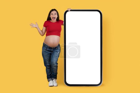 Foto de Gran Promo. Mujer embarazada joven asombrada parada cerca de un gran teléfono inteligente en blanco aislado sobre fondo amarillo, teléfono celular femenino expectante emocionado sorprendido con pantalla blanca, collage, burla - Imagen libre de derechos