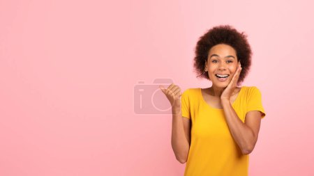 Photo for Cheerful surprised millennial african american curly woman in yellow t-shirt show finger at empty space, isolated on pink background, panorama, studio. Shocking offer and ad, huge sale and great news - Royalty Free Image