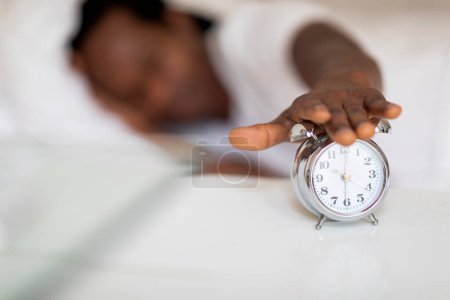 Foto de Primer plano de hombre afroamericano apagando el reloj de alarma con la mano, hombre negro irreconocible acostado en una cama cómoda en casa y llegando a la mesita de noche, despertando por la mañana, enfoque selectivo - Imagen libre de derechos