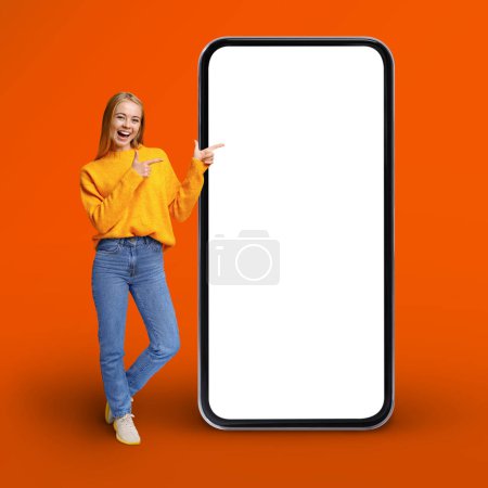 Photo for Great App. Positive Young Woman Pointing At Huge Blank Mobile Phone With White Screen While Standing Over Orange Background, Happy Cheerful Female Demonstrating Copy Space, Collage, Mockup - Royalty Free Image