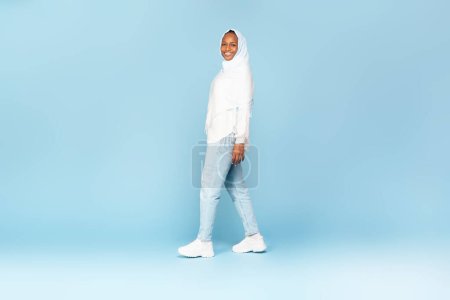 Téléchargez les photos : Heureuse femme afro-américaine en hijab marchant et souriant à la caméra, posant sur fond bleu studio, pleine longueur prise de vue. Offrir bannière publicitaire - en image libre de droit