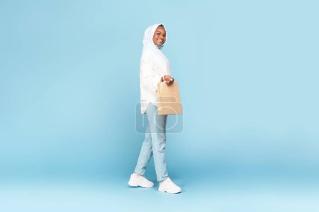 Photo for Seasonal sales concept. Full length shot of black muslim lady in hijab walking with shopping bag on blue studio background, free copy space - Royalty Free Image