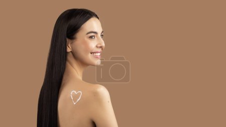 Photo for Beautiful brunette long-haired topless young lady with heart-shaped symbol on shoulder looking at copy space and smiling, enjoying nice beauty product, beige studio background, panorama - Royalty Free Image