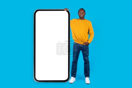 Photo for Cheerful cool adult black man in casual posing by huge smartphone with white empty screen on blue studio background, chatting with ladies via dating app, showing nice offer, mockup, full length - Royalty Free Image