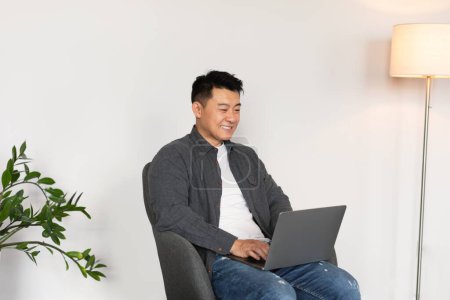 Foto de Feliz hombre japonés maduro escribiendo en el ordenador, ver vídeo, se sienta en sillón en el interior de la sala de estar con la pared blanca. Trabajo, negocios y freelance en casa, dispositivo moderno en la nueva normalidad durante covid-19 - Imagen libre de derechos