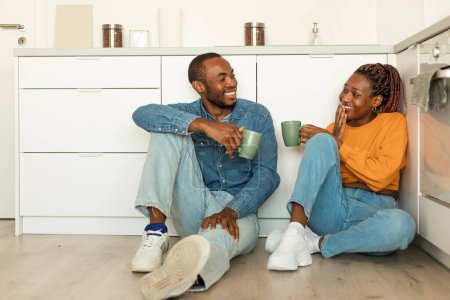 Téléchargez les photos : Couple afro-américain excité appréciant le café du matin à la maison, assis sur le sol dans la cuisine et ayant la conversation, tenant des tasses et bavardant, pleine longueur, espace libre - en image libre de droit