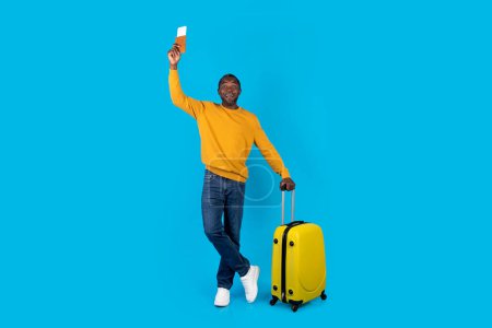 Photo for Cheerful african american middle aged man tourist travelling alone, carrying yellow luggage, showing passport with flight tickets, posing over blue studio background, copy space, full length - Royalty Free Image