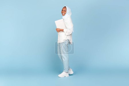 Téléchargez les photos : Heureuse femme afro-américaine en hijab tenant un ordinateur portable et marchant sur fond de studio bleu, pleine longueur prise de vue, espace libre. Internet business et freelance concept de carrière - en image libre de droit
