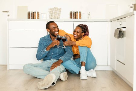 Foto de Romántica pareja afroamericana bebiendo vino tinto mientras está sentada en el suelo en la cocina, los cónyuges sosteniendo gafas y sonriéndose mutuamente, celebrando el aniversario en casa - Imagen libre de derechos