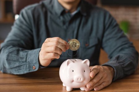 Reife asiatische männliche Manager legt Bitcoin-Münze in Sparschwein im Home Office Interieur, ausgeschnitten, aus nächster Nähe. Geld sparen, Investitionen in Kryptowährungen für Unternehmen und Arbeit, Start-up-Finanzierungen während covid-19