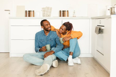 Photo for Loving black spouses bonding while drinking coffee, sitting on floor in kitchen, holding cups with hot drinks and enjoying spending time together at home, copy space - Royalty Free Image