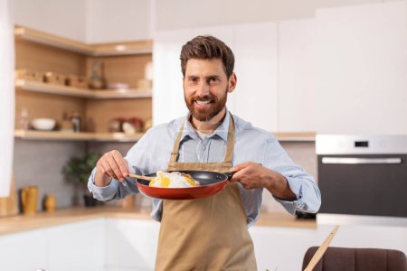 Photo for Happy attractive mature caucasian man with beard in apron prepare scrambled eggs in minimalist kitchen interior, free space. Breakfast in morning from chef, homemade eat, food blog and cook at home - Royalty Free Image