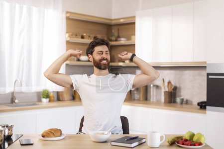 Photo for Glad cheerful adult caucasian man with beard in white t-shirt stretching his body in modern kitchen interior, free space. Guy enjoys good morning and breakfast at free time and weekend alone at home - Royalty Free Image