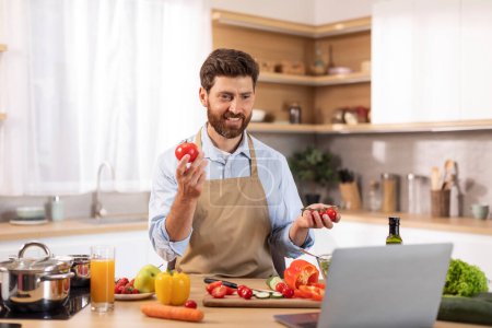 Photo for Happy mature caucasian male with beard in apron has video call, makes video holds tomato with hands in minimalist kitchen interior. Chef prepares healthy homemade food, meal blog with gadget at home - Royalty Free Image
