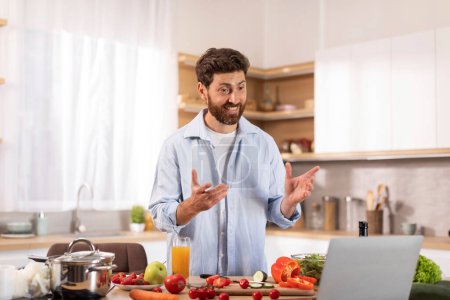 Photo for Cheerful middle aged caucasian man with beard has video call, make video for food blog, gesturing hands, preparing healthy meal in modern kitchen interior. Homemade eat, health care and device at home - Royalty Free Image
