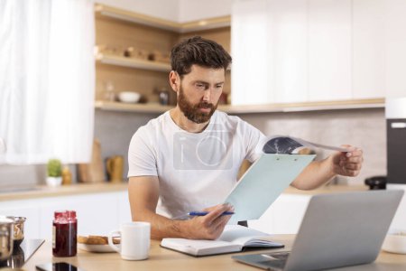 Photo for Serious adult caucasian male with beard works with documents and laptop in minimalist kitchen interior. Business, study and startup, online lesson and webinar at social distance with device at home - Royalty Free Image