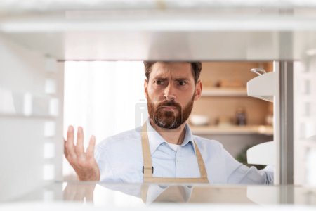 Photo for Sad hungry adult caucasian male with beard in apron opens door and looks into empty refrigerator in kitchen interior, free space. World crisis, starving, diet, problems in eating, no food at home - Royalty Free Image