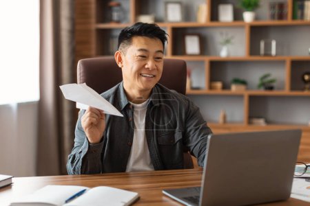 Foto de Sonriendo hombre asiático de mediana edad haciendo avión con papel, la estrategia de planificación en la mesa con el ordenador portátil en el interior de la oficina en casa. Creación de ideas para los negocios, lluvia de ideas, trabajo durante la pandemia de covid-19 - Imagen libre de derechos