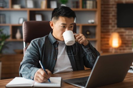 Téléchargez les photos : Occupé concentré d'âge moyen asiatique mâle boit du café, regarde l'ordinateur à la table, a appel vidéo à l'intérieur du bureau à la maison. Interrompre le travail, les affaires et le freelance à distance pendant la pandémie covid-19 - en image libre de droit