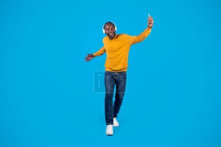 Photo for Positive energetic middle aged black man in stylish casual outfit with modern wireless stereo headphones and cell phone in his hand listening to music and dancing over blue, full length, copy space - Royalty Free Image