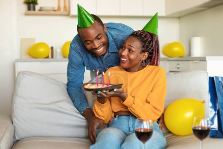 Foto de Cumpleaños de maridos. esposa afroamericana sorprendiendo a su marido con pastel de cumpleaños, hombre soplando velas y sonriendo, disfrutando de la celebración del día b en casa. - Imagen libre de derechos