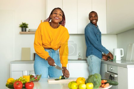 Foto de Alegre pareja negra cocinando comida y riendo, mujer preparando ensalada para la cena, de pie en la cocina moderna. Cónyuges afroamericanos cocinando comida. Nutrición saludable y concepto de recetas familiares - Imagen libre de derechos