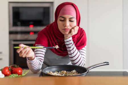 Foto de Mujer infeliz de Oriente Medio joven con hiyab musulmán ama de casa degustación de alimentos mientras cocina para su familia, marido en casa, insatisfecho con el resultado, interior de la cocina, espacio de copia. Escuela de cocina - Imagen libre de derechos