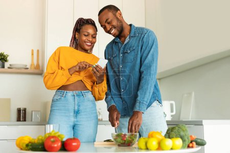 Téléchargez les photos : Épouses noires excitées en utilisant un téléphone portable tout en cuisinant le dîner ensemble et en naviguant sur Internet pour des recettes en ligne, debout dans la cuisine, espace libre. Joyeux couple préparant le repas à la maison - en image libre de droit