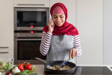 Téléchargez les photos : Conversation avec un ami au sujet de la cuisine. Jeune et jolie femme du Moyen-Orient portant le hijab parle sur un téléphone portable dans la cuisine tout en préparant le dîner tout en se tenant dans la cuisine - en image libre de droit