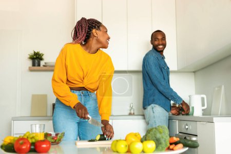 Foto de Feliz pareja afroamericana preparando comida saludable, pasando tiempo en la cocina juntos, hablando mientras cocinan y sonriendo el uno al otro, espacio libre - Imagen libre de derechos