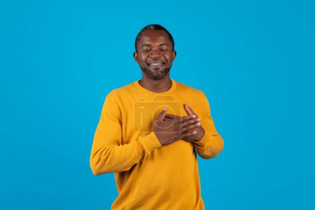 Téléchargez les photos : Paisible homme afro-américain d'âge moyen en jaune tenant la main sur sa poitrine au-dessus du cœur et souriant, priant les yeux fermés sur fond bleu studio, chérir l'espoir, copier l'espace - en image libre de droit