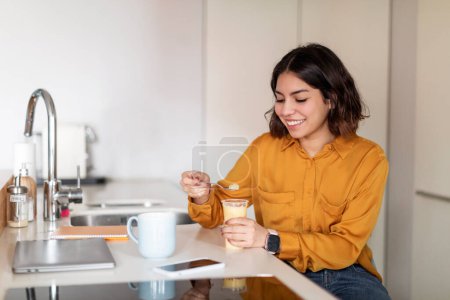 Photo for Young Arab Woman Eating Pudding And Drinking Coffee For Breakfast At Home, Happy Millennial Middle Eastern Female Sitting At Kitchen Counter And Enjoying Her Delicious Meal, Copy Space - Royalty Free Image