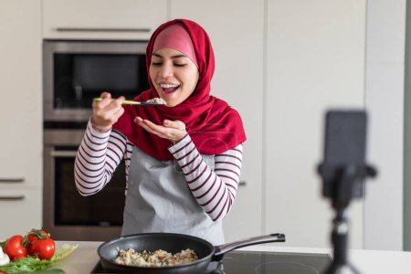 Téléchargez les photos : Vlog culinaire. Happy Islamic Woman In Hijab enregistrement vidéo tout en cuisinant dans la cuisine, Sourire musulmane dans le tablier en utilisant Smartphone sur le trépied à la maison, la capture de contenu pour les médias sociaux - en image libre de droit