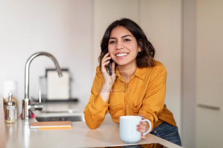 Téléchargez les photos : Souriante jeune femme arabe parlant sur son téléphone portable et buvant du café dans la cuisine à la maison, portrait d'une belle femme heureuse du Moyen-Orient ayant une conversation mobile agréable et profitant d'une boisson chaude - en image libre de droit