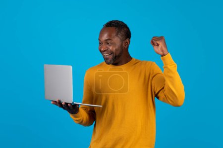 Foto de Emocional afroamericano hombre de mediana edad en suéter amarillo utilizando la computadora moderna en fondo de estudio azul, juegos de azar o comercio en línea, celebrando el éxito, apretando el puño y sonriendo, espacio de copia - Imagen libre de derechos