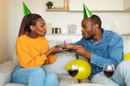 Foto de Pareja negra excitada en sombreros de fiesta soplando velas en la torta de cumpleaños y bebiendo vino, sentada en el sofá en casa. Feliz familia afroamericana joven celebrando b-day juntos en el interior - Imagen libre de derechos