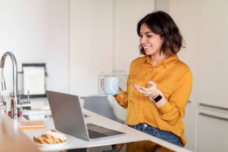 Téléchargez les photos : Femme arabe souriante faisant appel à la vidéo via un ordinateur portable et buvant du café dans la cuisine, Bonne jeune femme du Moyen-Orient parlant à la caméra Web de l'ordinateur, Profitant de la communication en ligne, Espace de copie - en image libre de droit