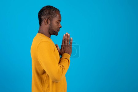 Photo for Side view of calm spiritual handsome mature black man praying with closed eyes. Serious peaceful man with joining hands meditating. Isolated on blue studio background. Belief concept, copy space - Royalty Free Image