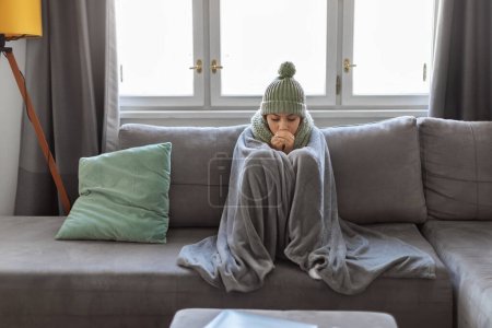 Téléchargez les photos : Jeune femme arabe souffrant de froid à la maison alors qu'elle était assise sur un canapé recouvert d'une couverture, femme millénaire portant une écharpe et un chapeau tricoté gelant dans le salon, ayant des problèmes de système de chauffage - en image libre de droit