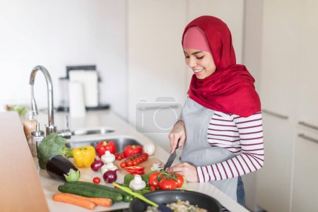 Foto de Agradable mujer joven de Oriente Medio vistiendo hijab rojo, delantal, cómodo traje casual ama de casa preparando la comida en casa, de pie junto a la mesa de la cocina, cortando diversas verduras. espacio de copia - Imagen libre de derechos