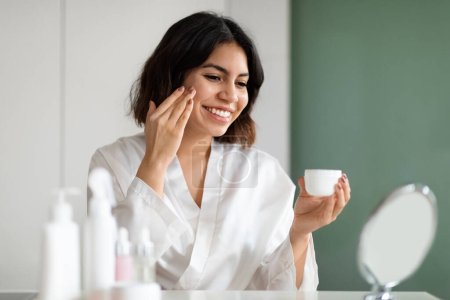 Skin Care Concept. Beautiful Female Using New Cosmetic At Home, Attractive Middle Eastern Young Woman In White Silk Robe Sitting In Front Of Mirror And Applying Moisturising Face Cream, Copy Space