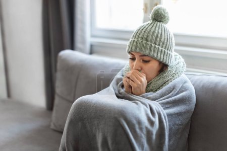 Photo for Young Woman Feeling Cold At Home, Freezing Female Sitting On Couch Covered In Warm Blanket, Millennial Lady Wearing Knitted Hat And Scarf Indoors, Warming Hands With Her Breath, Copy Space - Royalty Free Image