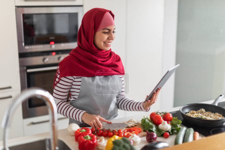 Téléchargez les photos : Heureuse belle jeune femme musulmane qui aime cuisiner à la maison, couper les légumes, lire la recette, blog alimentaire sur Internet, en utilisant une tablette numérique moderne tout en préparant un repas sain à la cuisine, copier l'espace - en image libre de droit